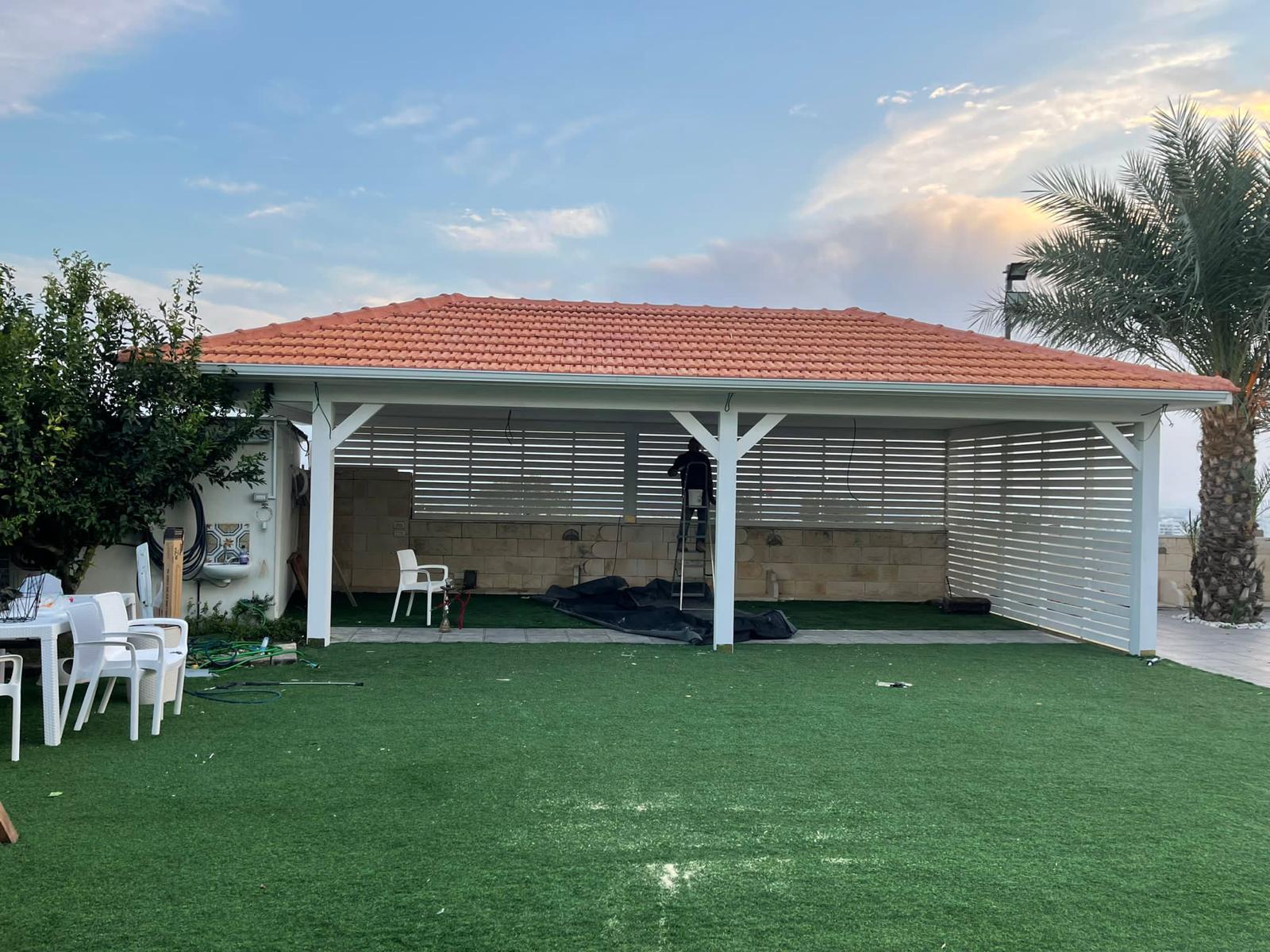 פרגולה לבנה עם קירוי רעפים כתום בחצר עם דשא